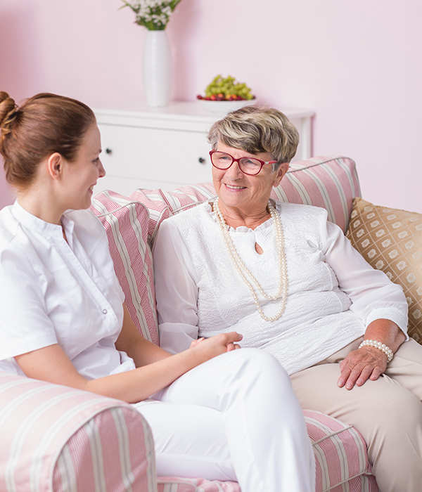 Caregiver and elderly woman smiling and talking while seated together on a couch,