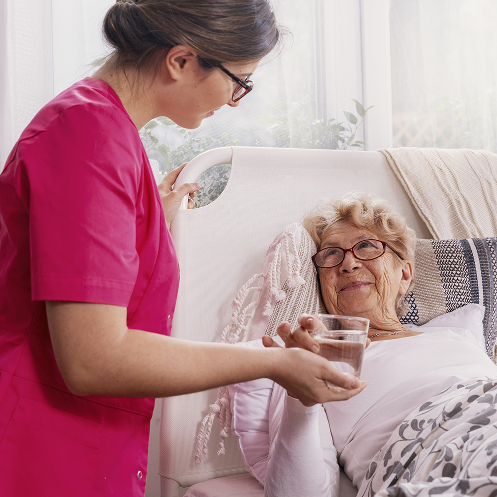 Nurse in pink scrubs helping an elderly woman in bed drink water, offering care and support.